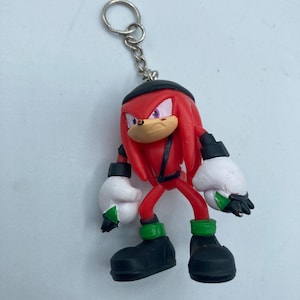 May include: A red Knuckles the Echidna keychain figurine. The figurine has white gloves with green accents, black shoes with green bands, and a black hat. The keychain is attached to a silver metal ring.