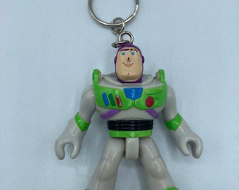 May include: A green and white plastic toy keychain of Buzz Lightyear from the Toy Story franchise. The keychain features a detailed depiction of Buzz Lightyear's iconic space ranger suit, complete with a purple helmet, green and blue accents, and a black belt.