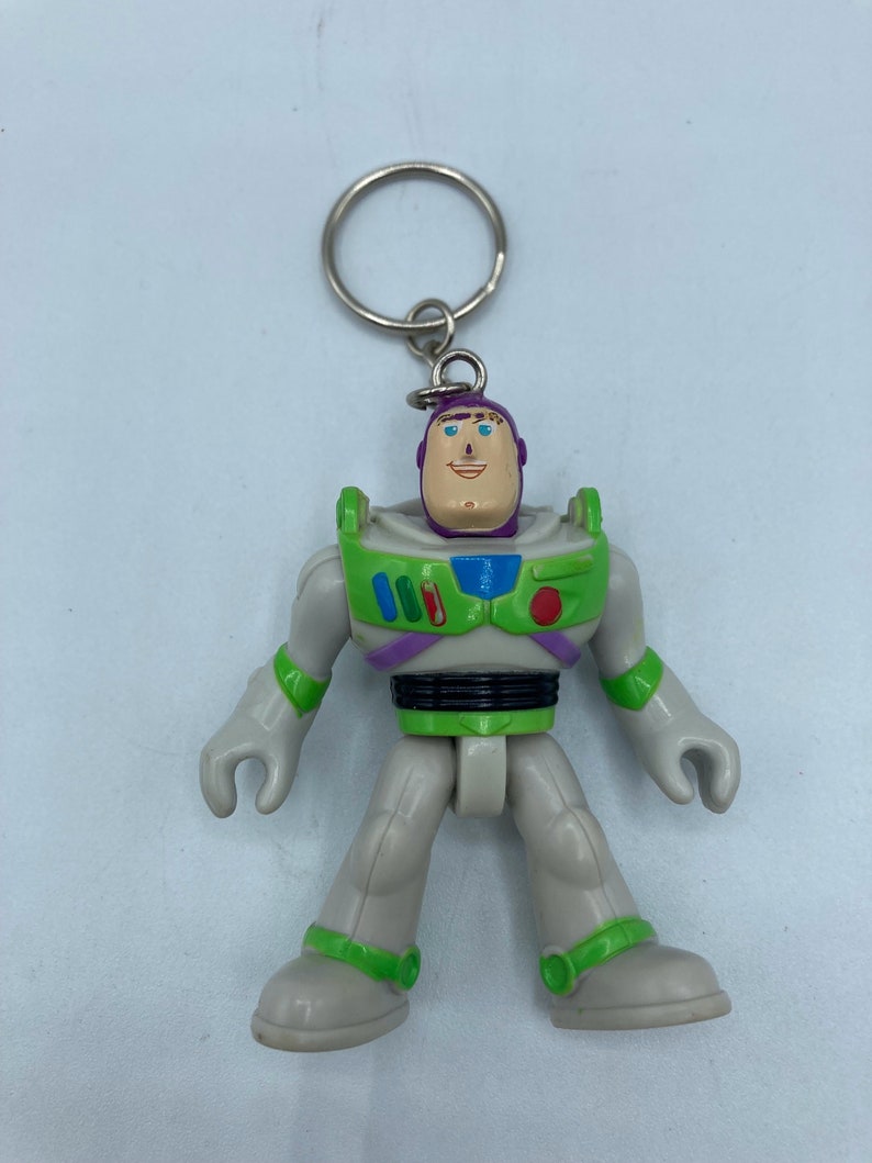May include: A green and white plastic toy keychain of Buzz Lightyear from the Toy Story franchise. The keychain features a detailed depiction of Buzz Lightyear's iconic space ranger suit, complete with a purple helmet, green and blue accents, and a black belt.