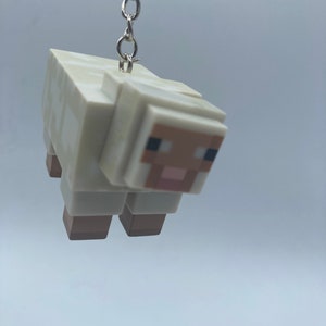 May include: A white and tan sheep keychain with a silver chain. The sheep has a blocky design, with a tan face and legs, and black eyes. The keychain is suspended against a light gray background.