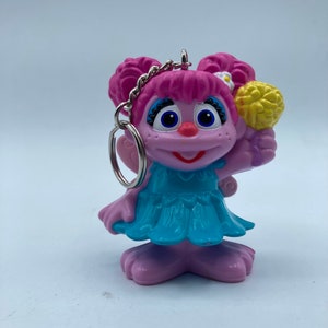May include: A pink and purple Sesame Street character keychain with a blue dress and a yellow flower. The character has pink hair and blue eyes.