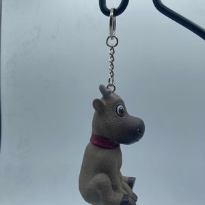 May include: A brown and gray reindeer keychain with a red collar. The reindeer is sitting with its head turned to the side.