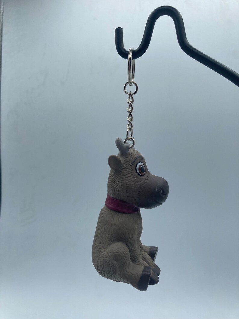 May include: A brown and gray reindeer keychain with a red collar. The reindeer is sitting with its head turned to the side.