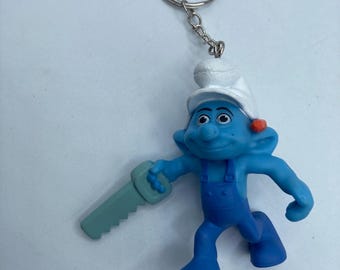 May include: A blue Smurf keychain featuring a Smurf character wearing blue overalls, a white hat, and holding a green saw. The keychain has a silver metal ring and chain. The Smurf has a cheerful expression.