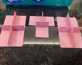 May include: Three pink, rectangular dividers with cross-shaped slots, designed for organizing items. The dividers are made of a solid, opaque material and are arranged on a dark, speckled surface. One divider is a single piece, while the other two are in two pieces.