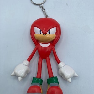 May include: A red Knuckles the Echidna keychain figure with white gloves, green and red shoes, and a silver keyring. The figure has a red body, a tan muzzle, and blue eyes. The keychain is a collectible item.