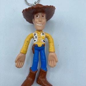 Pode incluir: Um chaveiro do Woody, personagem da franquia Toy Story. A figura tem um chap&eacute;u de cowboy marrom, camisa amarela, cal&ccedil;as azuis e botas marrons. O chaveiro tem um anel de metal prateado.