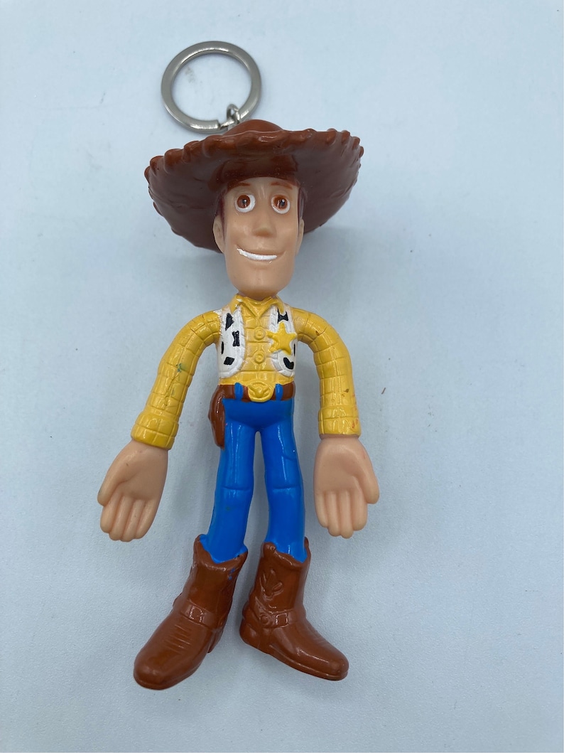 Pode incluir: Um chaveiro do Woody, personagem da franquia Toy Story. A figura tem um chap&eacute;u de cowboy marrom, camisa amarela, cal&ccedil;as azuis e botas marrons. O chaveiro tem um anel de metal prateado.