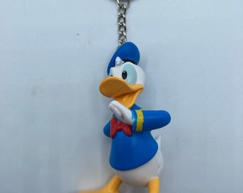 May include: A Donald Duck keychain featuring the iconic cartoon character in a blue sailor shirt, hat, and red bow tie. The keychain has a silver chain and ring. The figure is yellow, white, and blue.
