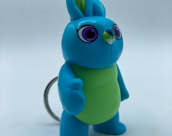 May include: A blue and green plush toy bunny keychain with purple eyes. The bunny is standing on a white surface.