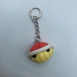 May include: A keychain featuring a cartoon character with a red shell, yellow face, and black eyes. The keychain has a silver chain and ring. The character's shell is red with a white border, and the face is yellow with black eyes.