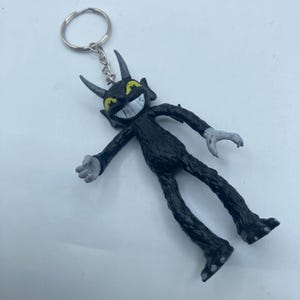 May include: Black devil-like keychain figurine with a silver keyring. The figurine has a wide grin, yellow eyes, and small horns. The arms and legs are outstretched, and the hands and feet are gray.
