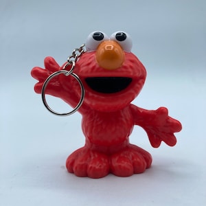 May include: A red Elmo keychain figurine with a silver keyring. The figurine has large white eyes, an orange nose, and a wide open mouth. The arms are outstretched in a welcoming gesture. The background is plain white.