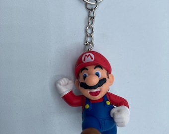 May include: A keychain featuring a small, colorful figurine of Mario, a popular video game character. Mario is depicted in his signature red hat with a white "M" emblem, red shirt, blue overalls, and brown shoes. The keychain has a silver metal ring and chain.
