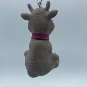 May include: A gray reindeer keychain with a pink collar. The keychain is made of plastic and has a metal chain attached to it. The reindeer is sitting down with its head tilted back. The keychain has the text "Disney" and "China" printed on the bottom.