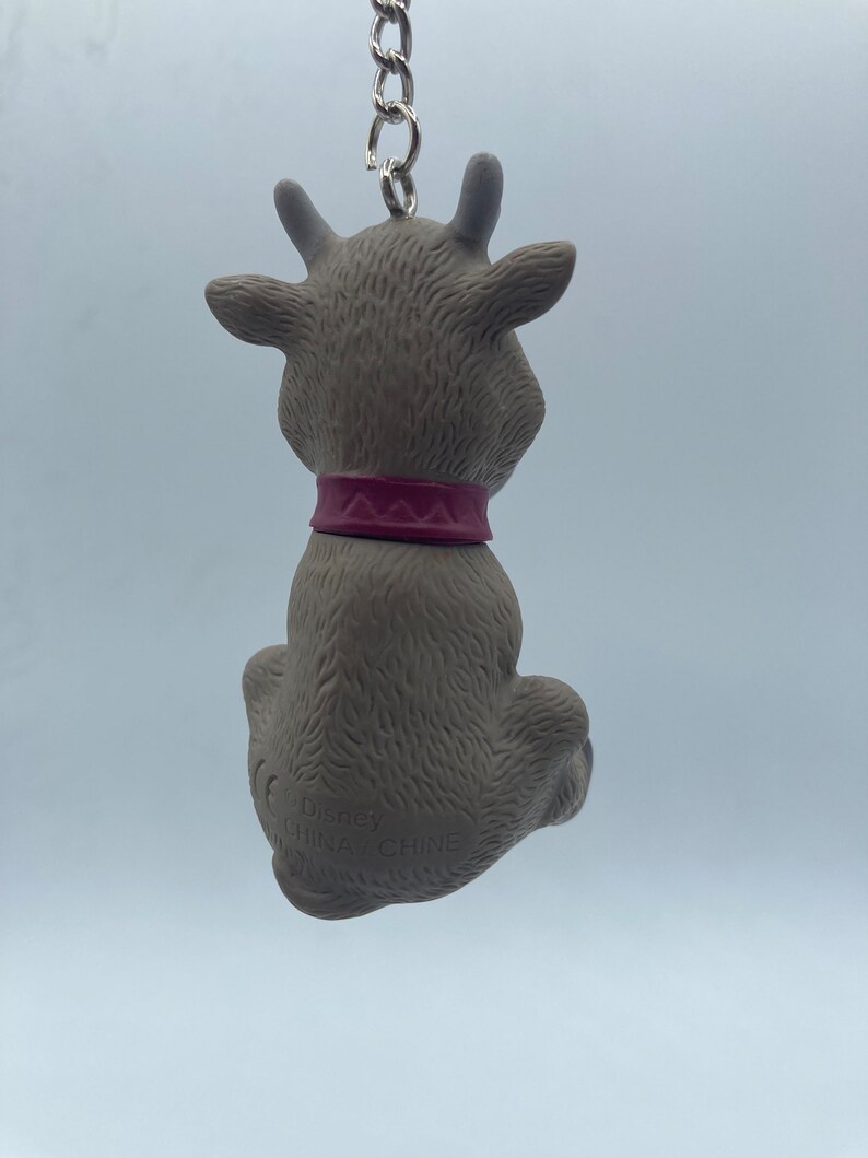 May include: A gray reindeer keychain with a pink collar. The keychain is made of plastic and has a metal chain attached to it. The reindeer is sitting down with its head tilted back. The keychain has the text "Disney" and "China" printed on the bottom.