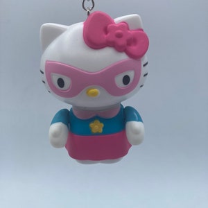 May include: A white Hello Kitty keychain with a pink bow, pink mask, and blue and pink superhero costume.