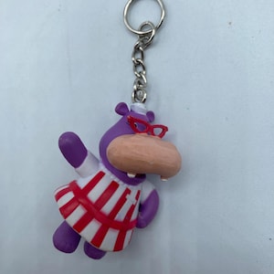 May include: A purple hippo keychain with a red and white striped dress, red glasses, and a light brown snout. The keychain has a silver chain and ring.