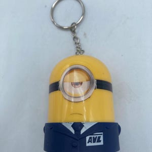 May include: A yellow Minion keychain with a silver key ring and chain. The Minion has one eye, black goggles, and a black band across the top. It is wearing a navy blue suit with the letters "AVL" on the front.