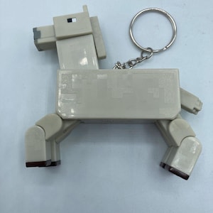 May include: A light beige horse-shaped keychain. The horse has a blocky design with dark brown hooves and a silver key ring. The keychain is made of plastic and has a pixelated appearance, resembling a character from a video game.