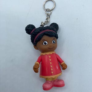 May include: A small, plastic keychain featuring a cartoon character. The figure has dark hair in two buns, wearing a red dress with tan trim and pink shoes. The keychain has a silver ring and chain.