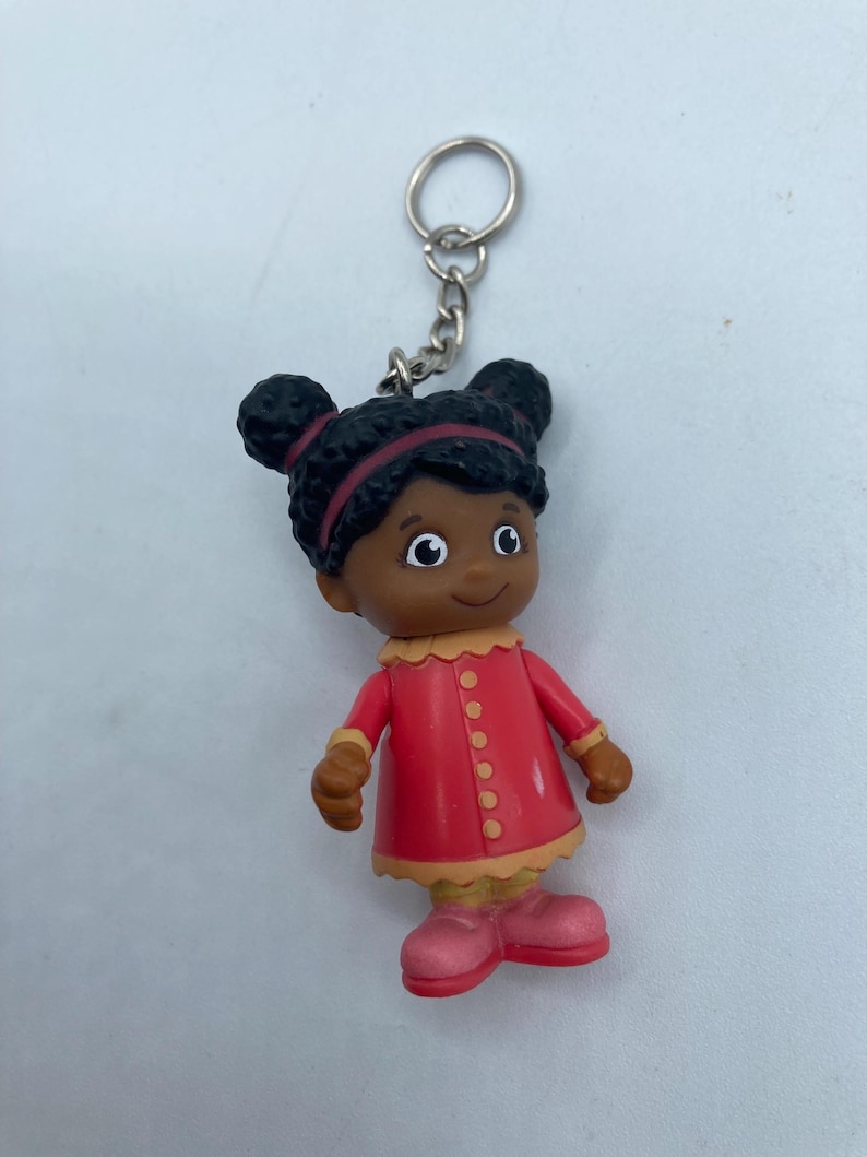 May include: A small, plastic keychain featuring a cartoon character. The figure has dark hair in two buns, wearing a red dress with tan trim and pink shoes. The keychain has a silver ring and chain.