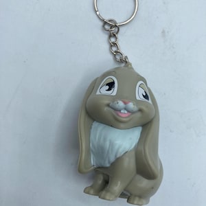 May include: A keychain featuring a cartoon bunny with long ears, a white chest, and a smiling face. The bunny is primarily tan with black and white accents. The keychain has a silver ring and chain.