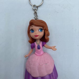 May include: A keychain featuring a small, plastic doll with brown hair and blue eyes. The doll wears a pink and purple dress and a silver necklace. The keychain has a silver ring attached to the top of the doll's head.