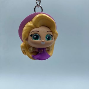 May include: A purple and pink keychain featuring a cartoon character with long blonde hair, a purple dress, and a pink hat. The character has large green eyes and a smiling face.