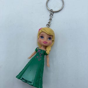 May include: A keychain featuring a doll with blonde hair and a green dress. The doll has a pink flower in her hair and is attached to a silver chain and keyring. The dress has pink floral accents.