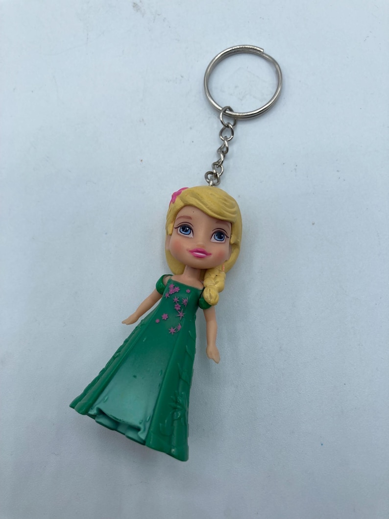 May include: A keychain featuring a doll with blonde hair and a green dress. The doll has a pink flower in her hair and is attached to a silver chain and keyring. The dress has pink floral accents.