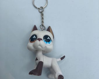 May include: A white and brown dog figurine keychain. The dog has large eyes with blue and brown irises, brown ears, and brown markings on its legs and tail. The keychain includes a silver metal ring and chain.