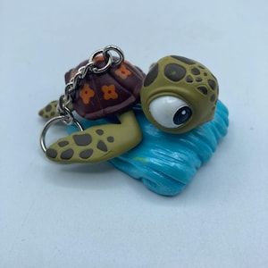 May include: A turtle-shaped keychain with a brown shell featuring orange flower designs. The turtle has a green head with dark spots and a large white eye. It rests on a blue wave-like base.