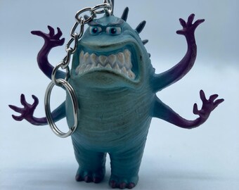 May include: A blue and teal monster keychain with a silver chain and ring. The monster has multiple arms with purple hands, a wide mouth with white teeth, and spiky details on its head. The keychain is a collectible item.
