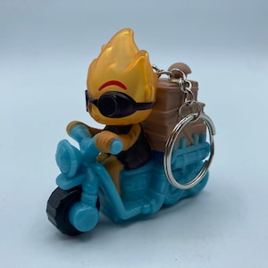 May include: A novelty keychain featuring a cartoon character with a flame-like head, wearing sunglasses, riding a light blue motorcycle. The character has a brown delivery box on the back of the motorcycle. The keychain has a silver ring.