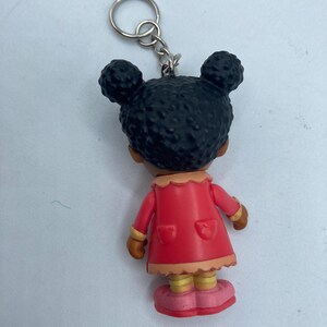 May include: A keychain featuring a small figurine of a girl with black hair styled in two buns. She wears a red coat with pockets, a tan collar, and pink shoes. The keychain has a silver ring.