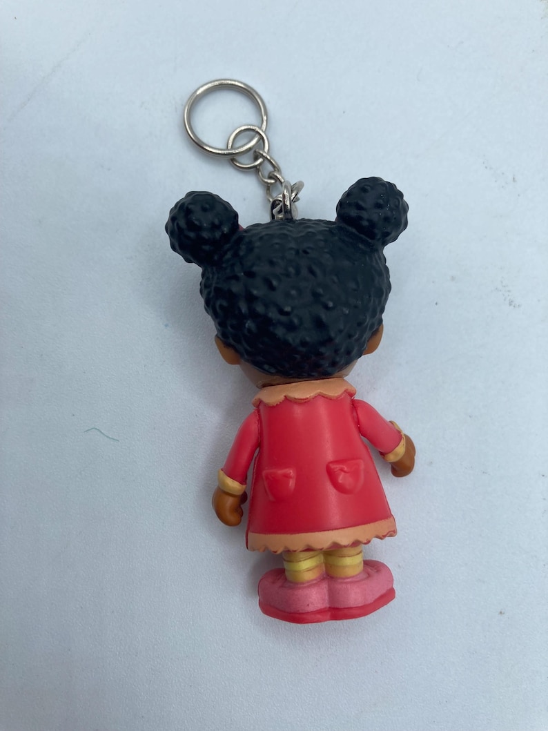 May include: A keychain featuring a small figurine of a girl with black hair styled in two buns. She wears a red coat with pockets, a tan collar, and pink shoes. The keychain has a silver ring.