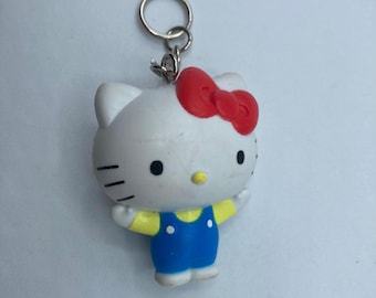 May include: A white Hello Kitty keychain with a red bow, black eyes, and a yellow nose. The figure wears a blue jumpsuit with yellow sleeves. A silver key ring is attached to the top.