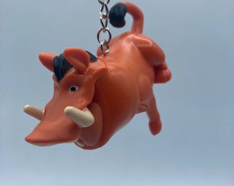 May include: A bright orange plastic keychain featuring Pumbaa from Disney's The Lion King. Pumbaa is a warthog with black eyes, a black snout, and white tusks. He is shown in a running pose with his tail curled up.