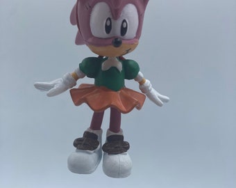 May include: A pink and white cartoon character with a green dress and orange skirt. The character is wearing white shoes with brown accents. The character is a keychain.