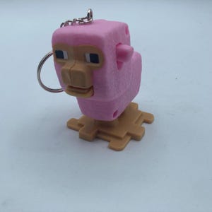 May include: A pink, pixelated pig keychain with a silver keyring and chain. The pig has a tan snout and feet, with black square eyes. The keychain is standing on a tan, pixelated base.