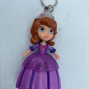 May include: A keychain featuring a small princess doll with auburn hair, blue eyes, and a purple dress. The doll has a silver tiara and is attached to a silver key ring. The dress has a glittery overlay.