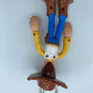 Pode incluir: Um chaveiro Woody, com chap&eacute;u de cowboy castanho, camisa amarela, cal&ccedil;as azuis e botas castanhas. A figura est&aacute; de cabe&ccedil;a para baixo, com um porta-chaves prateado preso ao chap&eacute;u.
