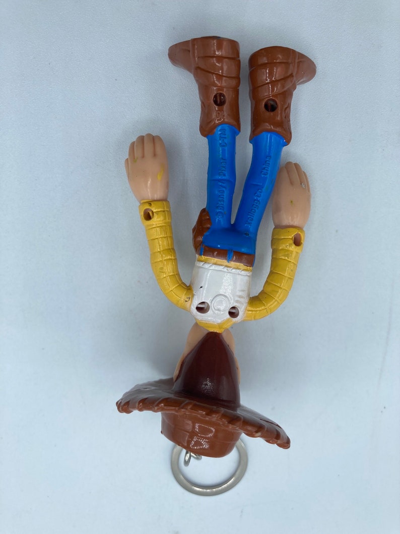 Pode incluir: Um chaveiro Woody, com chap&eacute;u de cowboy castanho, camisa amarela, cal&ccedil;as azuis e botas castanhas. A figura est&aacute; de cabe&ccedil;a para baixo, com um porta-chaves prateado preso ao chap&eacute;u.