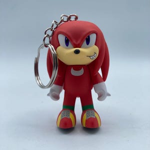 May include: A red Knuckles the Echidna keychain figure with a silver keyring. The figure has white hands, a tan muzzle, and yellow and red shoes. The figure is a character from the Sonic the Hedgehog video game series.