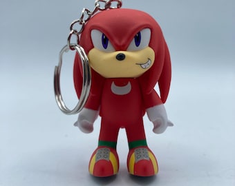 May include: A red Knuckles the Echidna keychain figure with a silver keyring. The figure has white hands, a tan muzzle, and yellow and red shoes. The figure is a character from the Sonic the Hedgehog video game series.