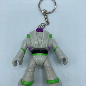 May include: A white and green Buzz Lightyear keychain with a purple dome on top. The keychain is attached to a silver key ring.