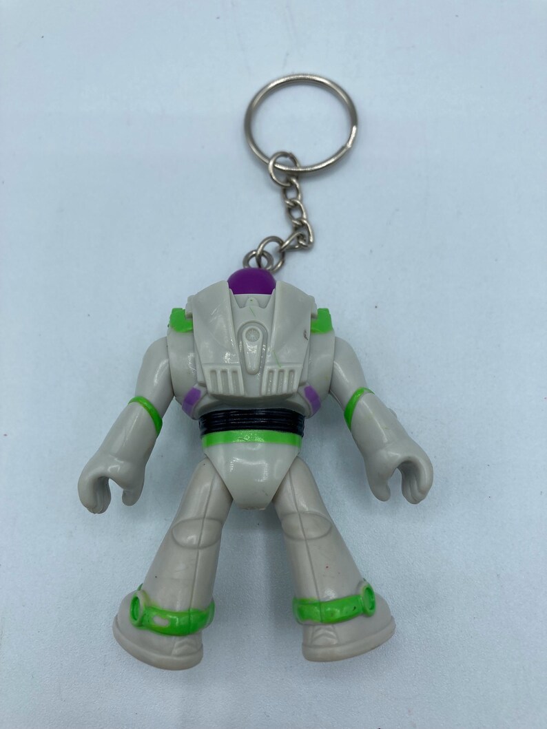 May include: A white and green Buzz Lightyear keychain with a purple dome on top. The keychain is attached to a silver key ring.