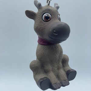 May include: A brown and gray reindeer keychain with large brown eyes and a purple collar. The reindeer is sitting with its legs crossed and has a silver chain attached to its back.