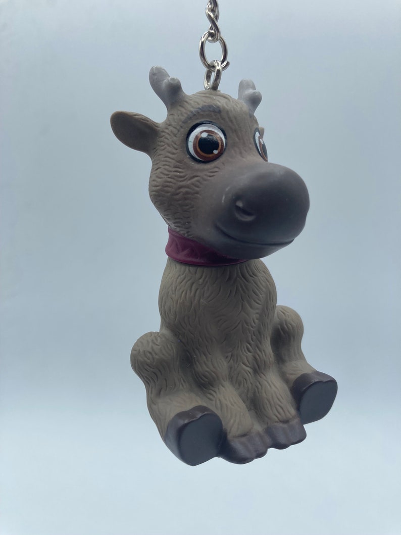 May include: A brown and gray reindeer keychain with large brown eyes and a purple collar. The reindeer is sitting with its legs crossed and has a silver chain attached to its back.